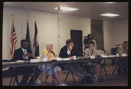 Five members seated behind a table, and a cameraman standing behind them. Featured left to right: Thaddeus Kirkland, E.Z. Taylor, Curt Schroder, Mary Ann Dailey, and John W. Fichter.