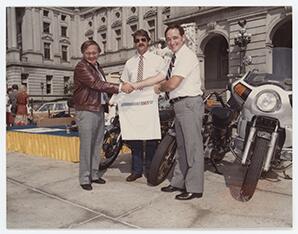 Rep. Brandt is holding one arm sleeve of a t-shirt and shaking hands with a member of the American Motorcycle Association