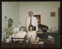 Rep. Burns in office standing behind Connie Clelan Betz, Secretary