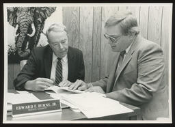 Rep. Burns speaks with a staff member at his desk. An elephant rug is in the background