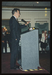 Campaign event, a man speaks at a podium, and Rep. Elinor Z. Taylor stands off in the background.